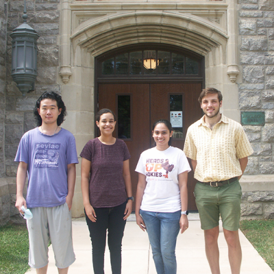 Four graduate students, stand in front of Davidson Hall. All students are facing the camera and smiling.