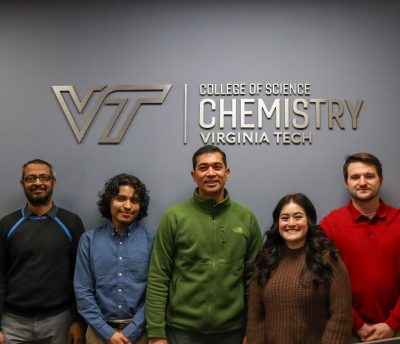 Five people stand against a wall with a sign that reads "Virginia Tech College of Science - Chemistry."